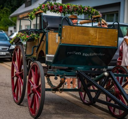 Pferdewagen mit blühender Dekoration, rote Räder, in einer Stadtstraße.