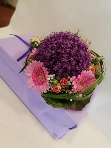 Runde Blumendekoration in Violett und Rosa mit grünen Blättern auf lila Unterlage.