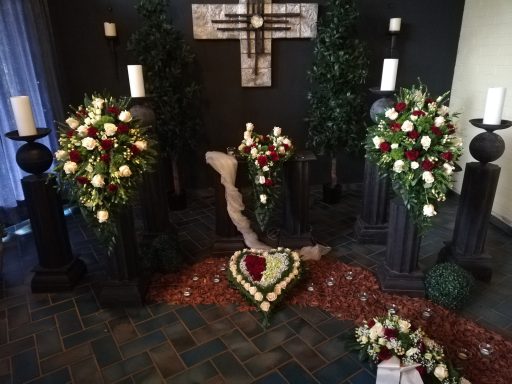 Trauerschmuck mit Blumen, Kerzen und Kreuz in einem Raum auf einem Teppich aus roten Steinen.