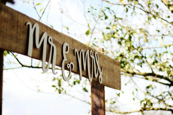 Holzschild mit der Aufschrift "Mr & Mrs" und grünen Blättern im Hintergrund.